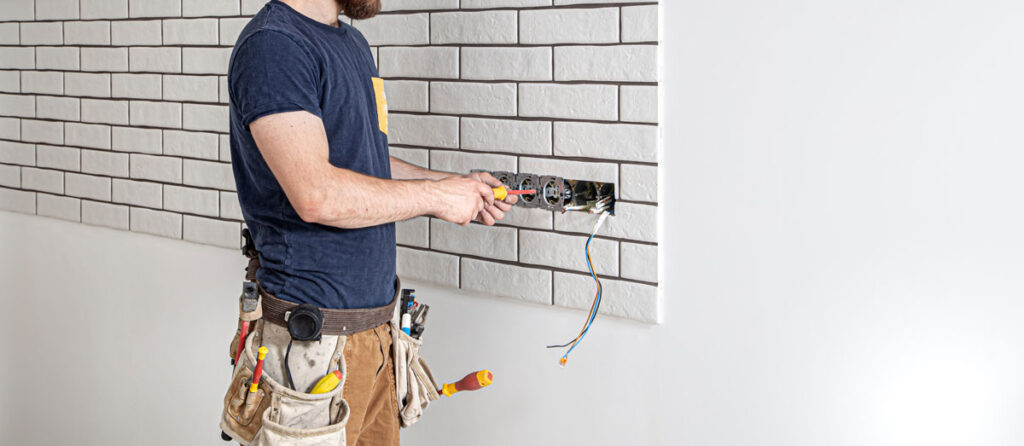 A licensed electrician with a tool belt works on an electrical wall box against white brick, handling the kind of same-day electrical repair ENS takes on.