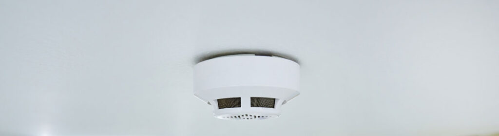A white smoke and carbon monoxide detector mounted on a ceiling, one of the safety devices ENS installs and tests during hardwired electrical work.
