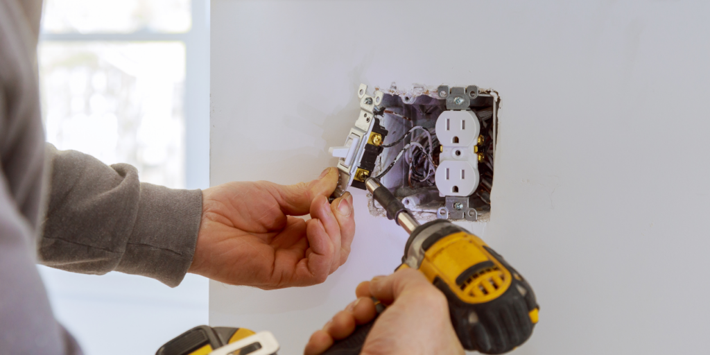 An ENS electrician drives a screw with a cordless drill while mounting a wall outlet, typical of the wiring and upgrade work done in older homes.