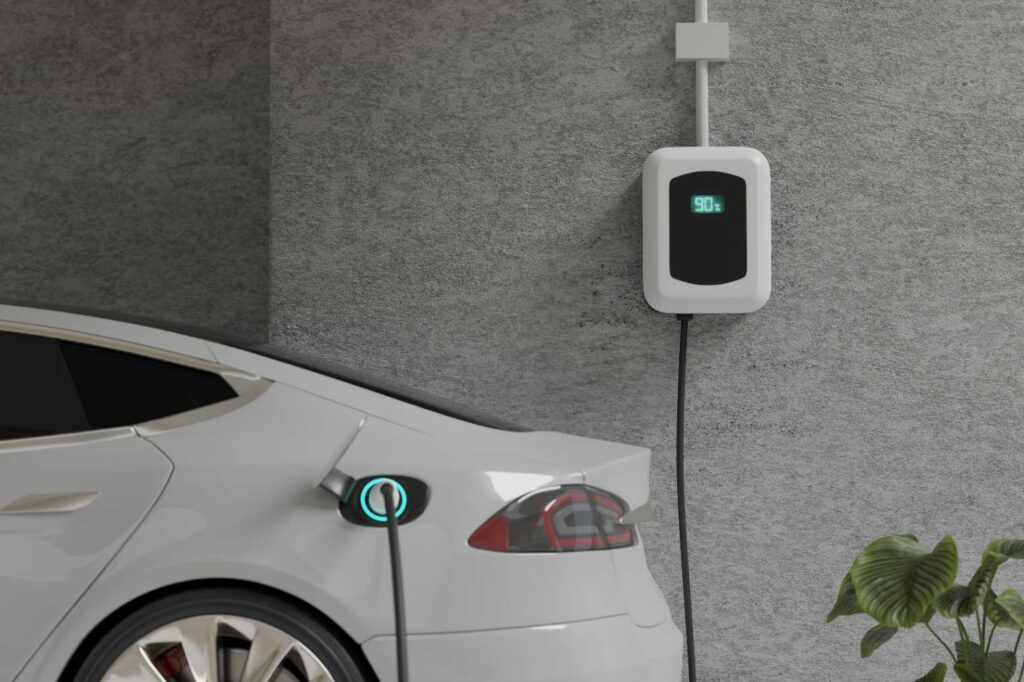 A Level 2 wall connector mounted on a garage wall with its cable plugged into a white car's charge port, wired to a dedicated 240V circuit.