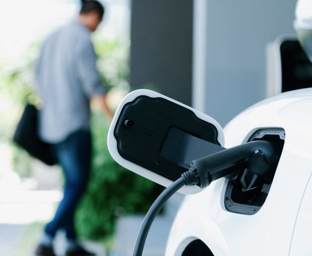 A charging connector plugged into a white EV car's port as the driver walks away, the everyday routine a home EV charger makes possible.