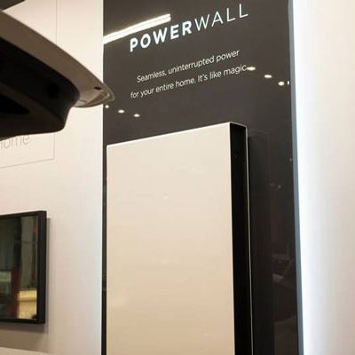 A Tesla Powerwall on display beside its signage describing seamless backup power for a whole home, one of the brands ENS carries and installs.
