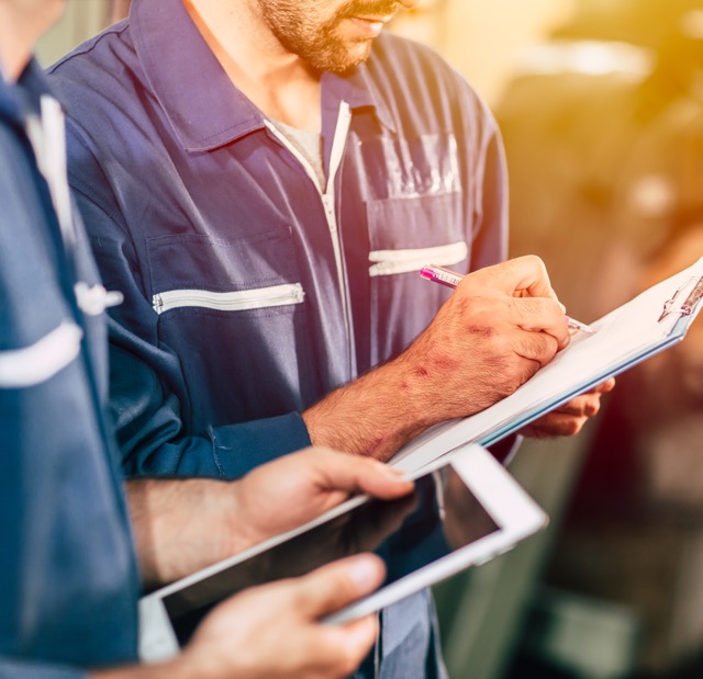 A generator technician in a blue work shirt writes service notes on a clipboard, part of the generator technician role open at ENS Electric.