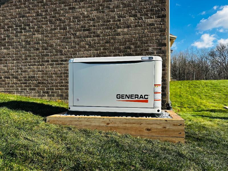 A white Generac standby generator, installed by ENS Electric, on a wood-framed gravel pad beside a brick home, ready to pick up the load when a storm drops the grid.