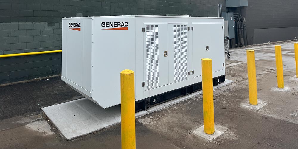 A large commercial Generac generator on a concrete pad protected by yellow bollards outside a West Michigan business that needs continuous power.